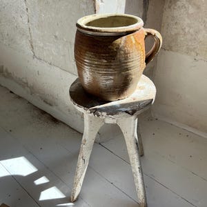 以下が含まれることがあります： 取っ手付きの素朴な茶色の陶器の壺が、小さな白い三本足のスツールの上に置かれています。壺は、テクスチャ加工されたリブのデザインと明るい色の内装が特徴です。スツールは、塗料が剥がれるなど、使用感があります。自然光で照らされたシーンです。