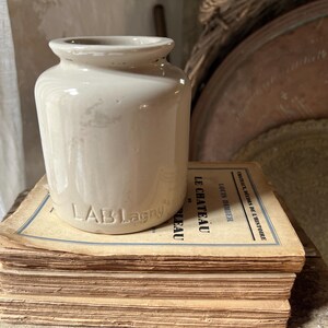 May include: A cream-coloured ceramic jar with the embossed text "LAB Lagny" sits atop a stack of aged books. The books have faded pages and French text. The jar has a cylindrical shape with a slightly flared rim.