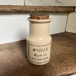 May include: A small, off-white ceramic jar with a cork stopper. The jar has a label with the text "MAILLE Moutarde à l'ancienne" in black script. The jar is sitting on a wooden surface.