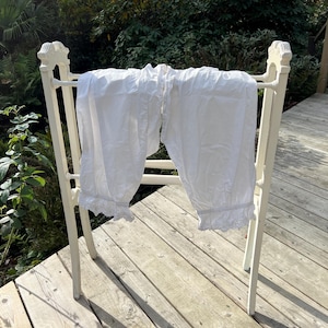 May include: White vintage bloomers with ruffled hems are draped over a white wooden drying rack. The rack has decorative finials and is positioned on a wooden deck. The background features lush green foliage.