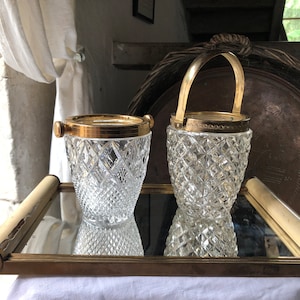 Op de afbeelding: Twee heldere, diamantgeslepen glazen ijsemmers met goudkleurige randen en accenten staan op een spiegelblad met gouden rand. Eén emmer heeft een handvat. De lade staat op een wit oppervlak.