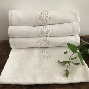 May include: Four white linen towels are stacked, each embroidered with a monogram. The towels are neatly folded, with the monogram visible on the top of each. A sprig of greenery rests on the wooden surface.