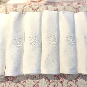 May include: Six rolled white linen napkins with embroidered details. Each napkin features a repeating geometric pattern and a stylized emblem. The napkins are arranged on a floral patterned surface.