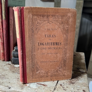 May include: A vintage book titled "TABLES DE LOGARITHMES A CINQ DECIMALES" by J. Dupuis. The cover is a faded brown with ornate detailing and red spine. Other books are visible in the background.