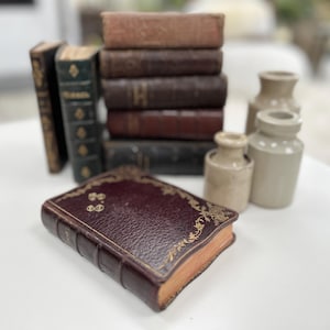 May include: A collection of antique books with leather-bound covers in various shades of brown and green. Some books are stacked, while others stand alone. Small, beige ceramic jars are also present, creating a vintage aesthetic.