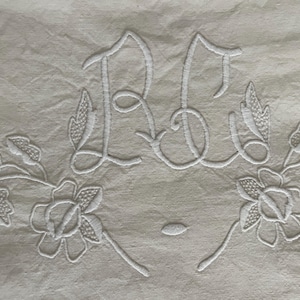 May include: Cream-colored textile with white embroidered initials "RB" intertwined with floral and leaf motifs. The embroidery is detailed, featuring delicate lines and shading, creating a textured appearance. The design is symmetrical, with flowers at the bottom.