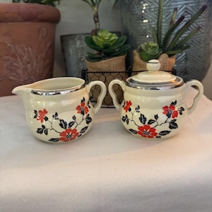 May include: Cream-colored creamer and sugar bowl set with red floral designs and silver trim. The sugar bowl has a lid and both pieces have handles. The set is on a white surface, with plants in the background.