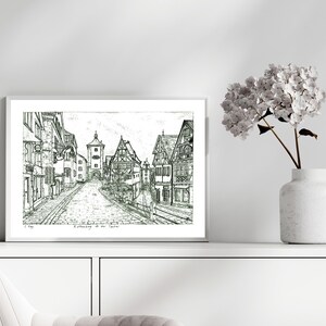 May include: Black and white ink drawing of a European town street scene, framed in a white border. The artwork depicts buildings, a tower, and a cobbled street. A white vase with flowers sits to the right.