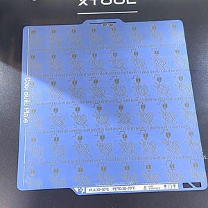 以下が含まれることがあります： 毛糸玉と編み針のアイコンが繰り返される模様が施された、正方形の青い3Dプリンター用ビルドプレート。左側に「Cool build plate」という文字があり、下端には「PLA:30-50°C PETG:50-70°C」と記載されています。