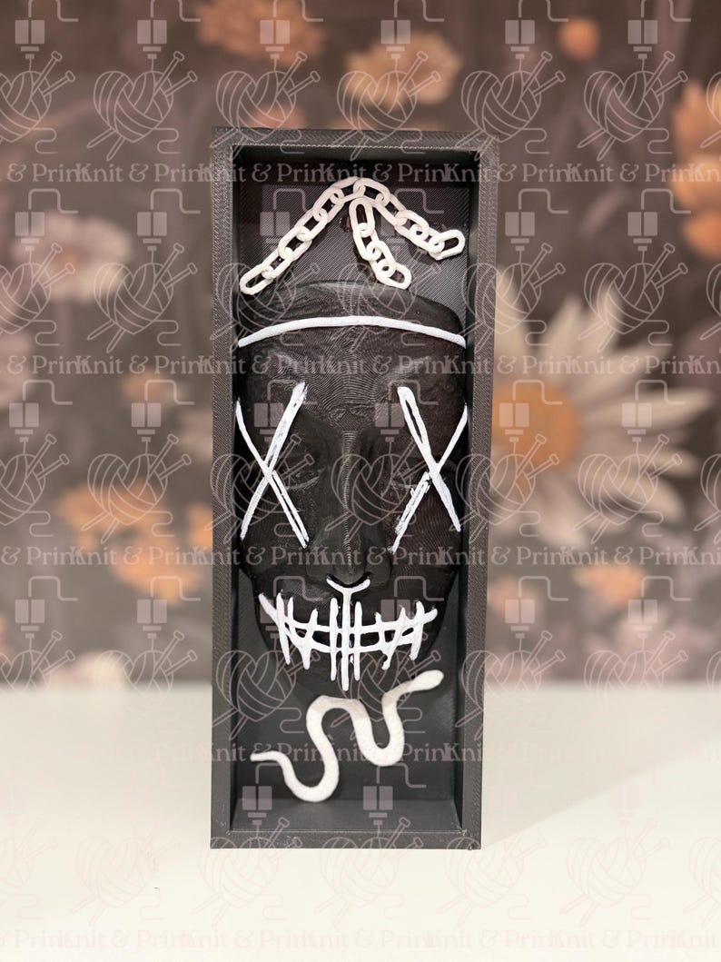 May include: A black mask with white accents in a rectangular frame. The mask features white X-shaped eyes, a stitched mouth, and a chain-like design above. The frame is dark gray, and the background has a repeating pattern.