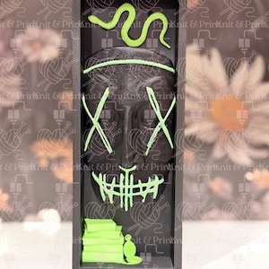 May include: A black rectangular display featuring a mask design with neon green accents. The mask has an X-shaped eye design and a mouth design. A neon green snake is at the top, and a stack of books and a bottle are at the bottom.