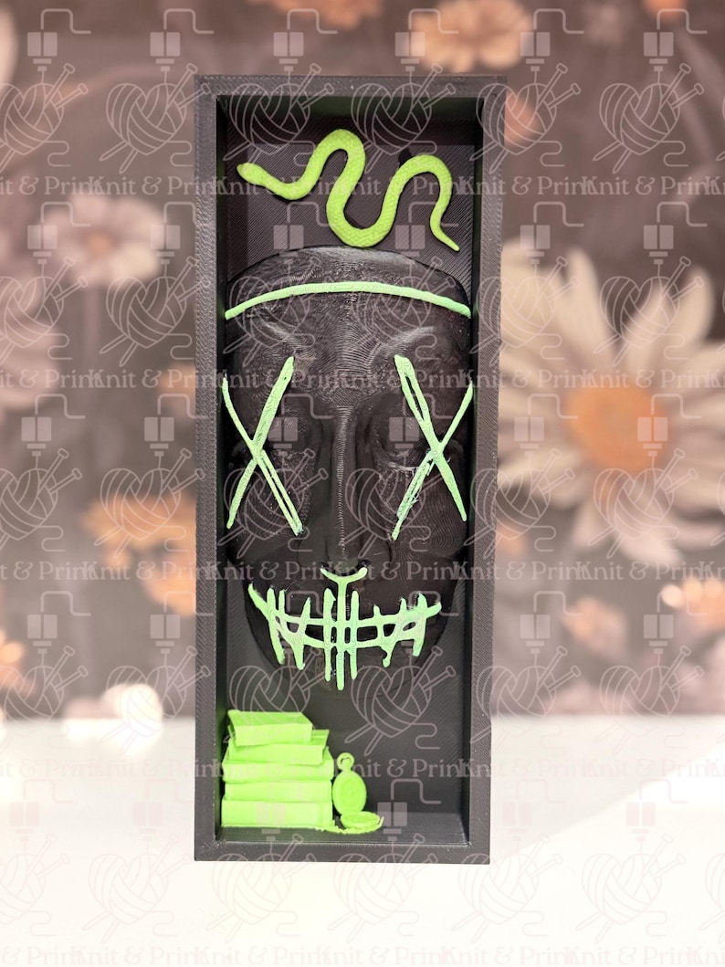 May include: A black rectangular display featuring a mask design with neon green accents. The mask has an X-shaped eye design and a mouth design. A neon green snake is at the top, and a stack of books and a bottle are at the bottom.