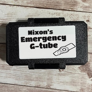 Könnte beinhalten: Ein schwarzer rechteckiger Koffer mit einem weißen Etikett mit der Aufschrift "Nixon's Emergency G-tube". Auf dem Etikett befindet sich eine Strichzeichnung eines medizinischen Geräts. Der Koffer befindet sich auf einer Holzoberfläche.