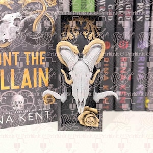 May include: A black bookend with a ram skull design, gold roses, and decorative gold accents. The bookend is positioned in front of a book with the title "UNT THE VILLAIN" and the author's name "NA KENT".