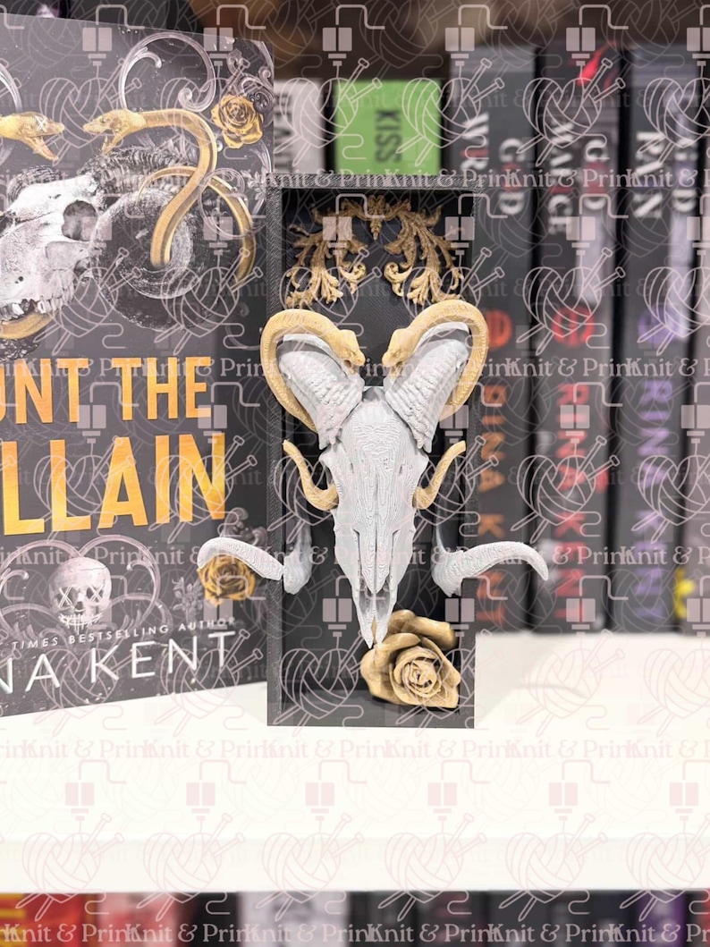 May include: A black bookend with a ram skull design, gold roses, and decorative gold accents. The bookend is positioned in front of a book with the title "UNT THE VILLAIN" and the author's name "NA KENT".