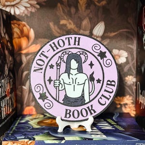 Puede incluir: Un posavasos redondo de madera de color morado y negro con el texto "Not-Hoth Book Club" y una ilustración en blanco y negro de una persona con cuernos y cola.