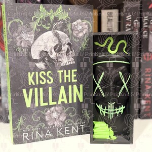 May include: A book titled "Kiss the Villain" by Rina Kent, with a skull and floral design on the cover. Beside it is a black box with a mask and a green snake design. The book and box are on a shelf.