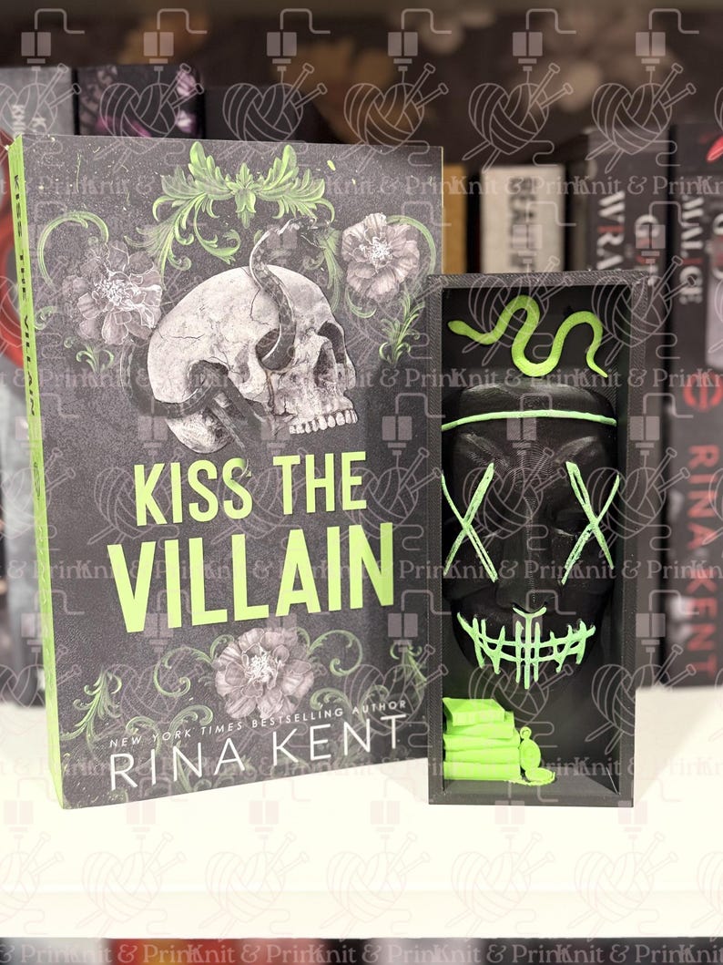May include: A book titled "Kiss the Villain" by Rina Kent, with a skull and floral design on the cover. Beside it is a black box with a mask and a green snake design. The book and box are on a shelf.