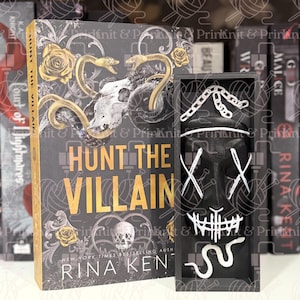 May include: A hardcover book titled "Hunt the Villain" with a skull and snake design, next to a black box containing a white mask with an "X" design. The book has gold accents.
