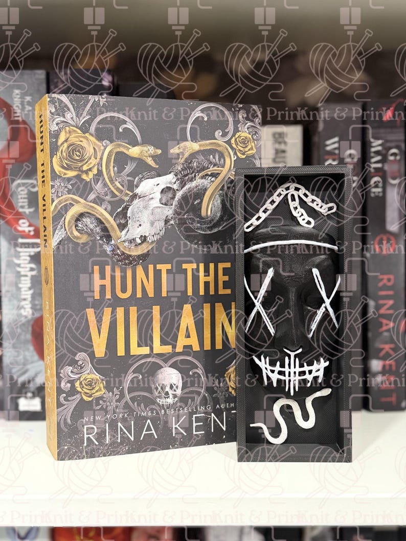 May include: A hardcover book titled "Hunt the Villain" with a skull and snake design, next to a black box containing a white mask with an "X" design. The book has gold accents.
