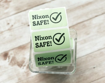 May include: Three green and white stickers with the text "Nixon SAFE!" and a black checkmark. The stickers are in a clear plastic dispenser.