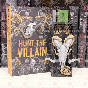 May include: A book titled "HUNT THE VILLAIN" with a skull and floral design, displayed with a matching skull-shaped bookend. The cover is black with gold lettering and accents. The author's name, Rina Kent, is visible. The book is on a shelf.
