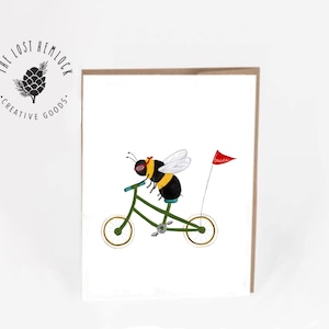 May include: A greeting card featuring a cartoon bumblebee riding a green bicycle with a red flag that says "Buzz" on it. The card is white with a brown border.