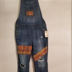 May include: Blue denim overalls with distressed details and colorful embroidered patches. The overalls have a relaxed fit and a straight leg.