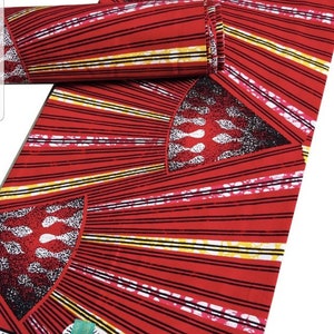 May include: Red fabric with a repeating pattern of black and yellow stripes radiating from a central design. The fabric has a rolled section and a flat section. The design includes white and black shapes. The fabric has the text "VERITABLE WAX ANDAIS VLISCO".