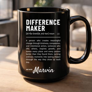 May include: A black ceramic mug featuring white text that defines the term Difference Maker as a person who creates meaningful change through kindness and compassion, followed by the text See also: Marvin, with a phonetic pronunciation guide.