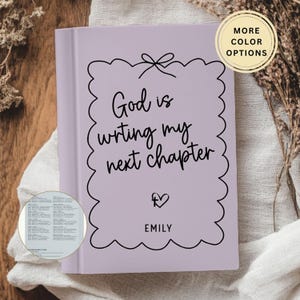 May include: A lavender journal with the text "God is writing my next chapter" in black script. The journal has a scalloped border and a small heart with the name "Emily" below. A circular button indicates "More Color Options".