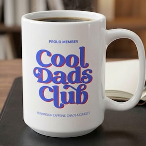 May include: White ceramic coffee mug filled with dark liquid, featuring the text "Cool Dads Club" in blue with a red shadow. The mug also has the text "Proud Member" and "Running on Caffeine, Chaos & Cuddles".