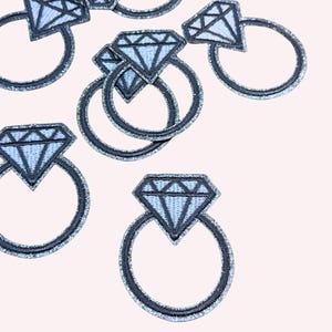 May include: Embroidered diamond ring patches. Each patch features a light blue diamond and a dark grey ring, with glittery thread detailing. The patches are scattered on a pale pink background.