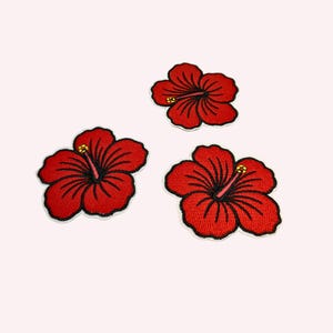 May include: Three embroidered hibiscus flower patches featuring vibrant red petals with black stitched detailing and a central yellow stamen. Each flower has five rounded petals and a distinct outline, presented as individual fabric appliqués.