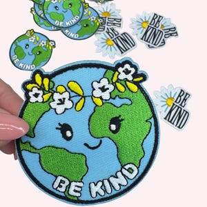 May include: Embroidered patches featuring a smiling Earth with a floral crown and the words "BE KIND". The patches are blue, green, yellow, and white. Additional patches with daisies and the words "BE KIND" are also visible.
