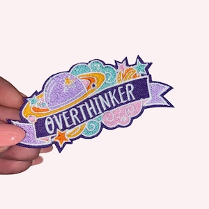 May include: Embroidered patch with the word "OVERTHINKER" on a purple banner. The patch features a purple planet, orange and yellow rings, teal clouds, pink swirls, and stars. The patch is held by a person.