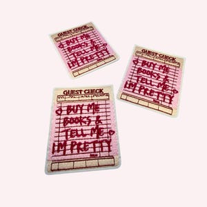 May include: Three rectangular embroidered patches shaped like guest checks. Each patch features a light pink background with dark red stitched text that reads: BUY ME BOOKS & TELL ME I'M PRETTY, along with small heart icons and grid lines.