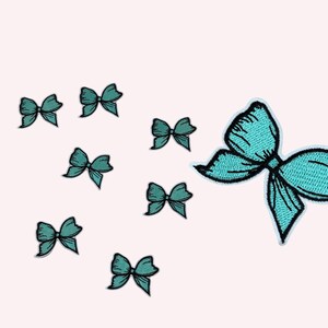 May include: A collection of teal bow patches with black outlines. The patches vary in size, with one large bow patch and several smaller ones scattered across a light pink background. The bows are embroidered.
