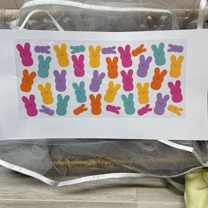Op de afbeelding: Een vel kleurrijke Paashaasstickers in verschillende kleuren, waaronder roze, blauw, geel, oranje en paars. De stickers zijn gerangschikt op een witte achtergrond, met een mesh-tas op de achtergrond.