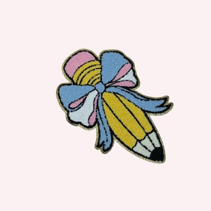 Puede incluir: Un parche colorido con forma de lápiz y un lazo. El lápiz es amarillo con detalles en blanco y negro, y el lazo presenta secciones rosas, azules y blancas. El parche tiene un contorno dorado brillante sobre un fondo rosa claro.