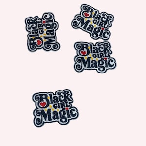 May include: Four embroidered patches with the words "Black Girl Magic" in a stylized font. The patches are black with white trim, a red heart, and yellow stars. The patches are arranged on a light pink background.