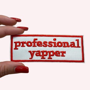 Könnte beinhalten: Ein rechteckiger, weißer Aufnäher mit rotem Rand und den Worten "professional yapper" in Rot gestickt. Der Aufnäher wird zwischen Fingern mit rotem Nagellack gehalten.