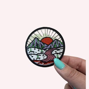 May include: Round embroidered patch featuring a mountain landscape with a red sun, purple mountains, and a pink river. The patch has a black border and is decorated with green trees and white accents.