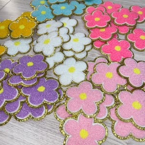 May include: A collection of colorful flower-shaped patches in various shades, including yellow, white, blue, pink, and purple. Each patch has a gold glitter outline and a yellow center, creating a vibrant and decorative effect.