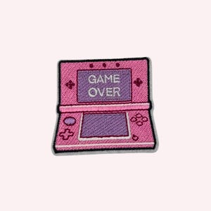 May include: An embroidered patch shaped like a handheld video game console in shades of pink and purple. The top screen displays the white text GAME OVER. The device features a directional pad, buttons, and two screens with a black border outline.
