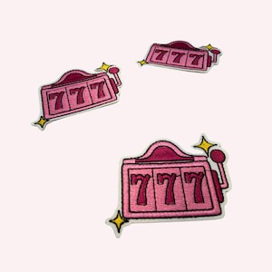 May include: Three embroidered patches shaped like slot machines in shades of pink and dark pink. Each patch features the number 777 in dark pink, a side lever, and small yellow star accents at the corners. The designs are outlined in white thread.