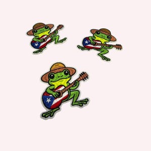 Puede incluir: Tres parches bordados que muestran una rana verde con un sombrero marrón tocando una guitarra decorada con el diseño de la bandera de Puerto Rico, que incluye un triángulo azul, una estrella blanca y franjas horizontales rojas y blancas.