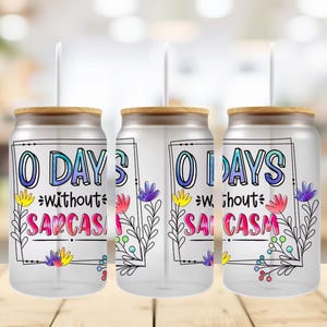 May include: Three clear glass can-shaped tumblers with bamboo lids and white straws. Each tumbler features the text "0 DAYS without SARCASM" in colorful lettering, framed by floral illustrations.