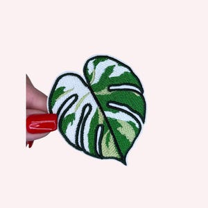 May include: Embroidered leaf patch featuring a variegated green and white Monstera design with black outlining. The patch is held against a light background, showcasing the detailed stitching and vibrant colors.