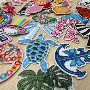 May include: A collection of embroidered patches in various shapes and colors. Designs include a blue turtle, a butterfly, a bow, a peach with the word "peachy", and a peace sign with flowers. Some patches have text, such as "I'M IN MY Goose Era."